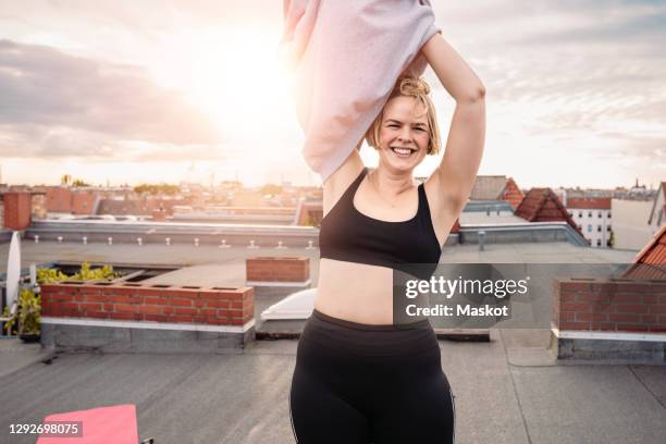 portrait of smiling woman removing t-shirt on rooftop against dramatic sky - mit nehmen stock-fotos und bilder