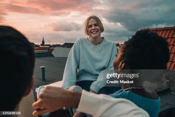 portrait of smiling female sitting with friends on building terrace during sunset - begrippen en thema's stockfoto's en -beelden