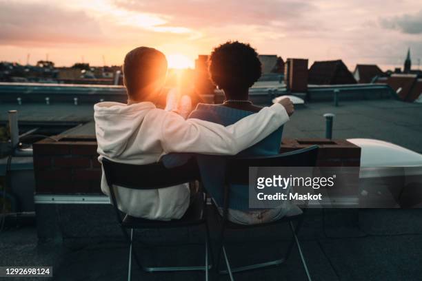 rear view of man and woman sitting on building rooftop during sunset - sunset stock-fotos und bilder