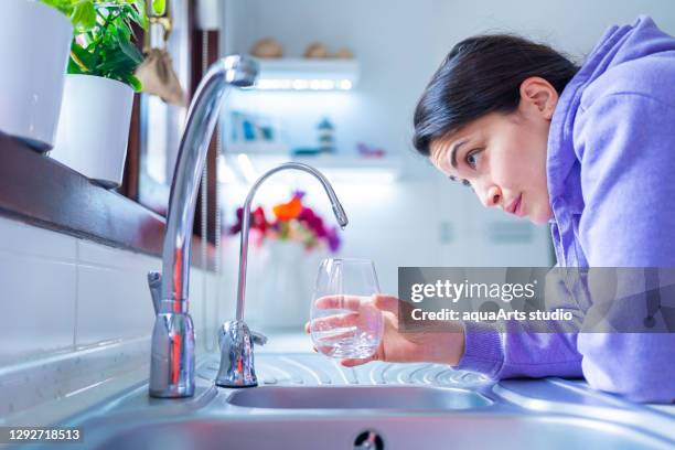 young and worried woman is waiting for a last drop of water hangs from a tap with glass under water shortage struck by drought .water shortage and drought crisis with global warming - faucet imagens e fotografias de stock