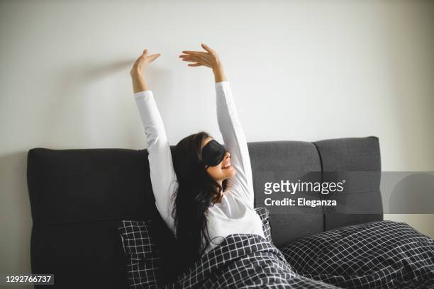 young woman wearing sleeping mask and stretching while sitting in her bed - máscara de olhos imagens e fotografias de stock