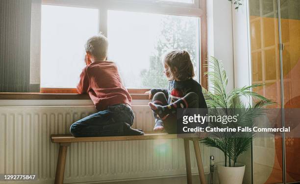 young girl and boy sit on a bench by a sunny window and gaze out - calor fotografías e imágenes de stock