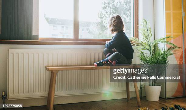 little girl sitting on a bench by a sunny window in a domestic room, looking out of the window - quarantine stock pictures, royalty-free photos & images