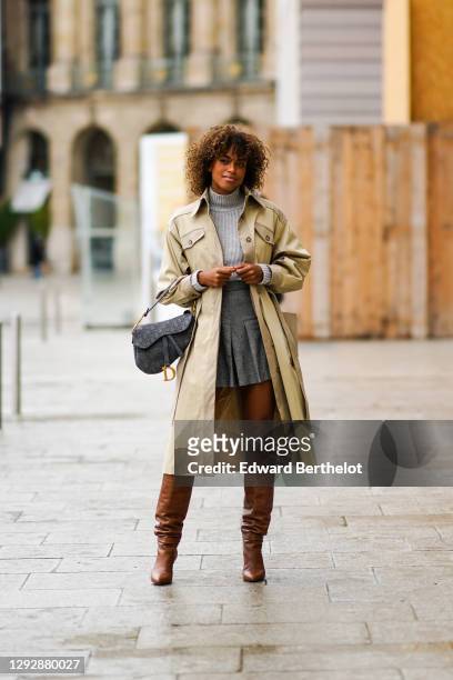Guest wears a gray wool turtleneck pullover, a beige long trench coat, a gray pleated short skirt, a Dior monogram Saddle bag, brown knee high...