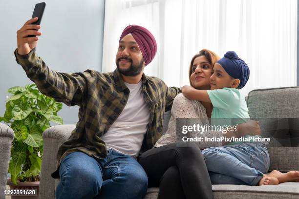 a sikh son and mother hug each other while father clicks a selfie of everyone sitting together on a sofa - father clicking selfie stock pictures, royalty-free photos & images