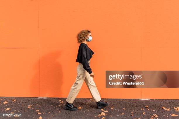 young woman wearing face mask walking against orange wall - protective face mask side stock pictures, royalty-free photos & images