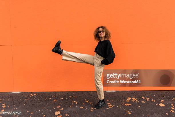 carefree woman wearing sunglasses stretching leg while standing on footpath - cool attitude stockfoto's en -beelden