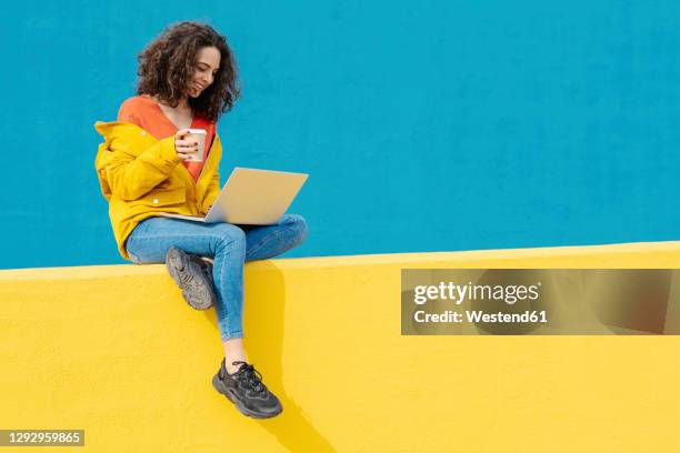 happy young woman with coffee to go sitting on yellow wall using laptop - leisure work coffee happy stockfoto's en -beelden