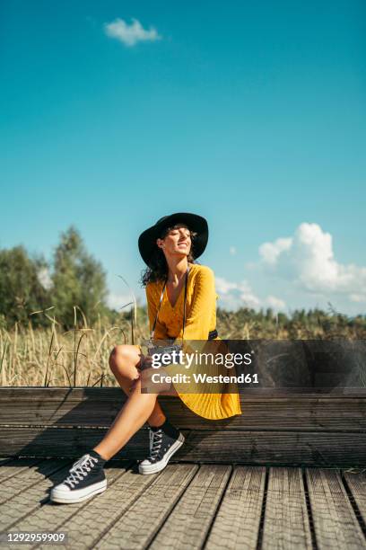young woman wearing a black hat and yellow dress with an analog camera sitting on wooden boardwalk - zapatos-amarillos fotografías e imágenes de stock