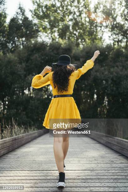 rear view of a young woman wearing black hat and yellow dress enjoying while is walking along wooden bridge - robe jaune photos et images de collection