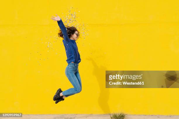 young woman jumping in the air in front of yellow background throwing confetti - celebration fotografías e imágenes de stock