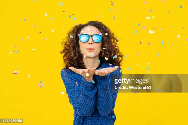 portrait of young woman with mirrored sunglasses blowing confetti in the air in front of yellow background - leichter stock-fotos und bilder
