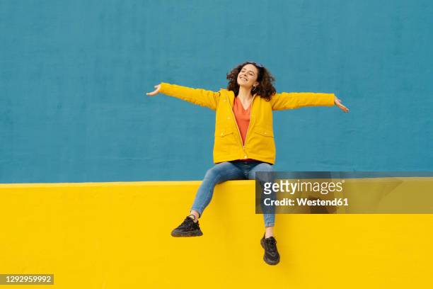 happy young woman sitting on yellow wall with arms outstretched - euforie stockfoto's en -beelden