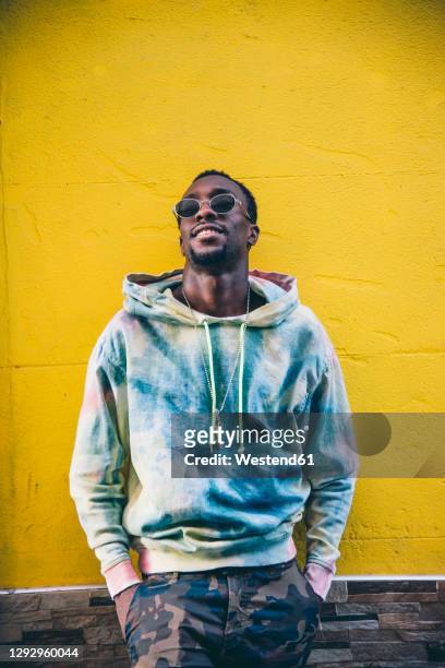 portrait of young man with sunglasses leaning against yelow wall - menswear stock-fotos und bilder