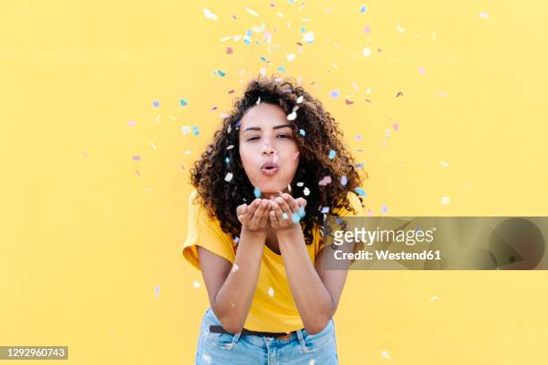 young woman blowing multi colored confetti against yellow wall - confetti stockfoto's en -beelden