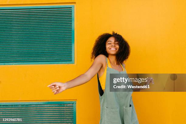 portrait of happy young woman wearing overalls in front of yellow wall - dans kleur stockfoto's en -beelden