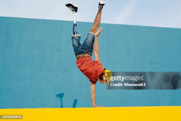 young man with artificial limb and foot doing handstand against multi colored wall - disabled athlete foto e immagini stock