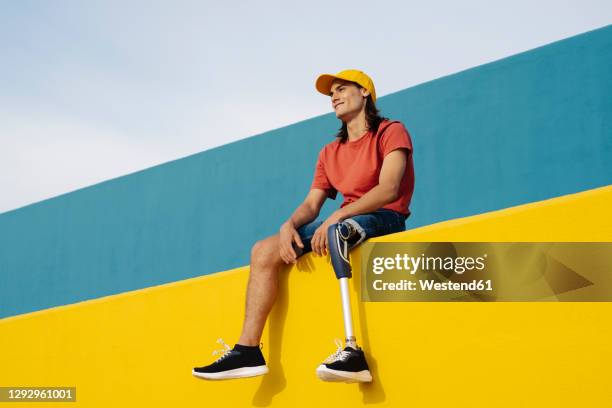 young man smiling while sitting on wall against clear sky - prosthetic stock-fotos und bilder