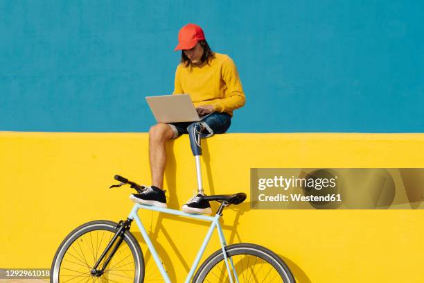 man with artificial limb and foot working on laptop while sitting on multi colored wall - laptop sparse stock pictures, royalty-free photos & images