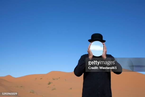 morocco, merzouga, erg chebbi, man wearing a bowler hat holding mirror in front of his face in desert dune - invisible man fotografías e imágenes de stock