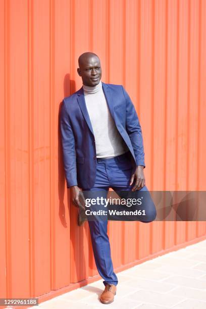 portrait of businessman wearing blue suit and grey turtleneck pullover leaning against orange wall - human knee 個照片及圖片檔