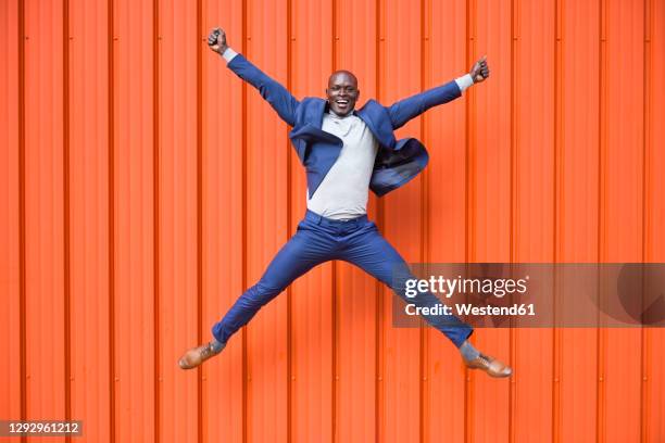 happy businessman jumping in the air in front of orange wall - jumping for joy stockfoto's en -beelden