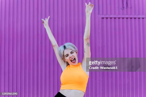 beautiful blond woman standing in front of purple wall, raising arms, laughing - braccia alzate foto e immagini stock