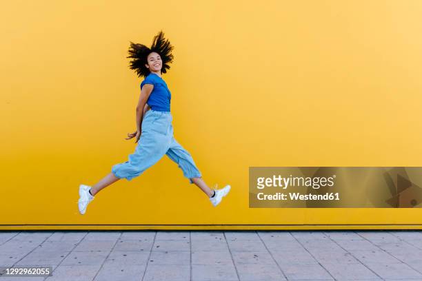 pretty woman jumping for joy in front of yellow wall - colore brillante foto e immagini stock