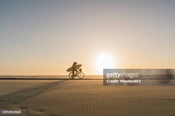 surfer riding a bicycle during the sunset in the beach, costa nova, portugal - bike beach stockfoto's en -beelden