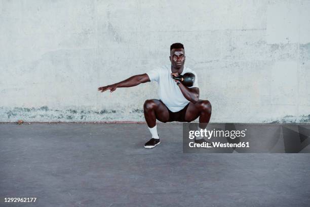 sportsman with arms outstretched holding kettle bell while crouching against wall - kettle bells stock pictures, royalty-free photos & images