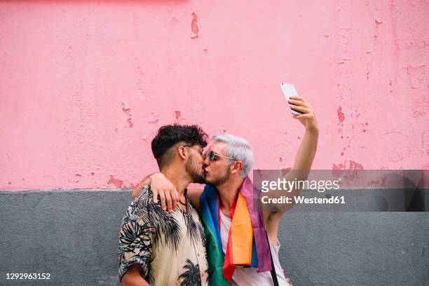 gay couple kissing and taking a selfie in front of pink and grey wall - gay couple kissing fotografías e imágenes de stock