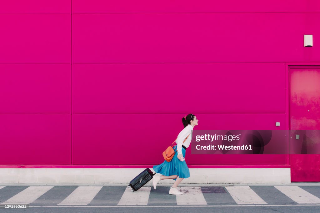 Young woman with smartphone running with trolley along a pink wall