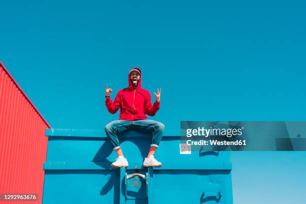 young man wearing red hoodie, sitting on edge of blue container, making sign of the horns - cool attitude foto e immagini stock