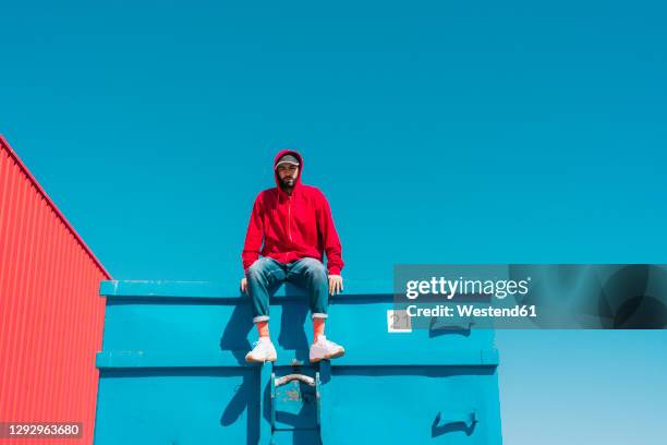 young man wearing red hoodie, sitting on edge of blue container - young man in attitude stock pictures, royalty-free photos & images