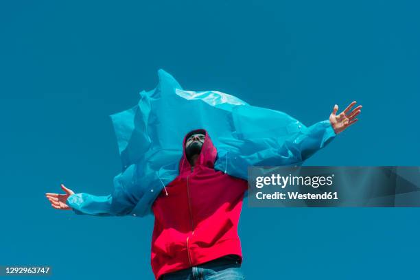 young man wearing plastic rain coat, stanind in gthe wind with arms outstretched - wolkenloser himmel stock-fotos und bilder