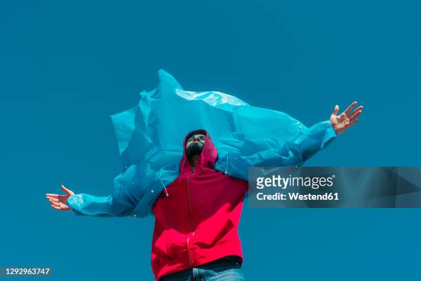 young man wearing plastic rain coat, stanind in gthe wind with arms outstretched - multi colored jacket stock pictures, royalty-free photos & images