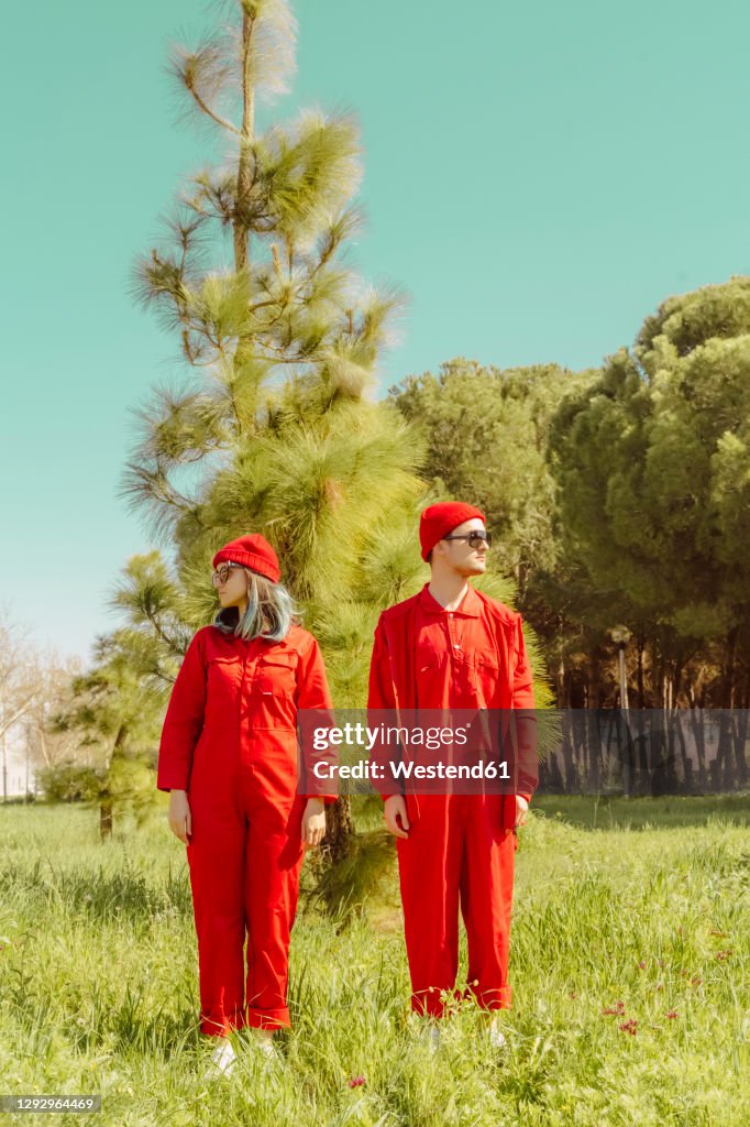 Young couple wearing red overalls and hats standing on a meadow looking around
