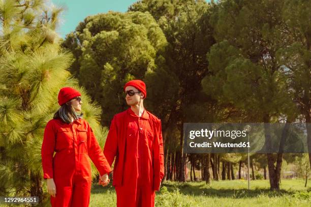 young couple wearing red overalls and hats standing hand in hand on a meadow looking at each other - matching outfits stock pictures, royalty-free photos & images