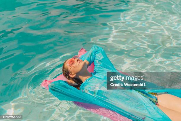 woman wearing blue rain coat relaxing on pink airbed in swimming pool - plastic pool stockfoto's en -beelden