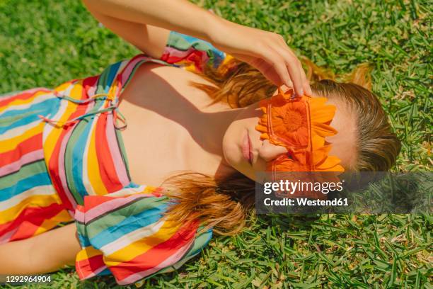 portrait of woman lying on a meadow wearing glasses with orange flowers covering her eyes - multi colored dress - fotografias e filmes do acervo