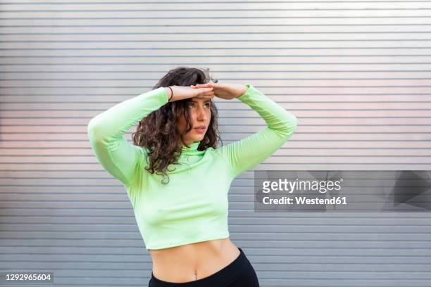 young woman shielding eyes while posing against gray wall in city - bauchfreies oberteil stock-fotos und bilder