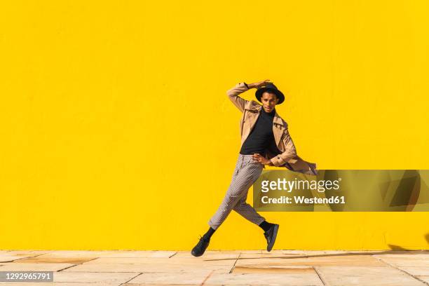 young man dancing in front of yellow wall, jumping mid air - fashion stock-fotos und bilder