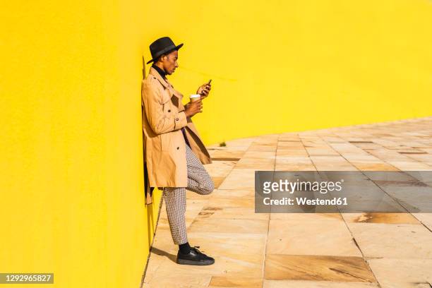 young man with hat, using smartphone, drinking coffee - trench stock photos et images de collection
