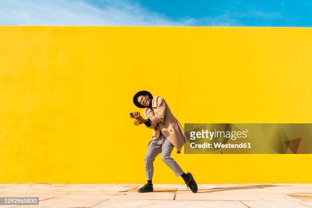 young man dancing in front of yellow wall, taking selfies - trench stock photos et images de collection