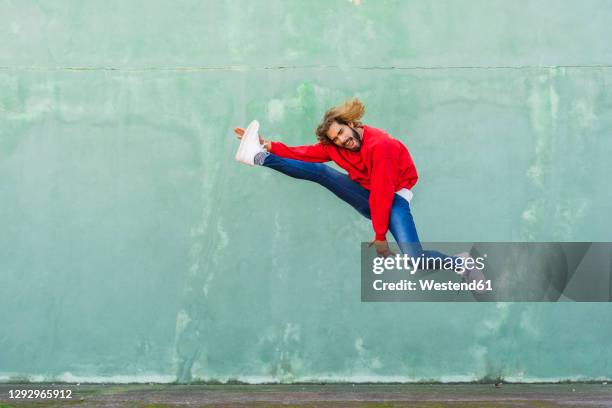 portrait of young man in wearing red sweatshirt jumping in the air in front of green wall - reaching higher stock pictures, royalty-free photos & images