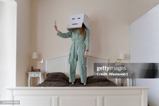 woman wearing a cardbox on head with bored smiley taking a selfie on her bed - irony stock pictures, royalty-free photos & images