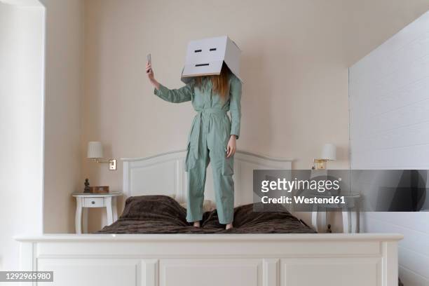 woman wearing a cardbox on head with bored smiley taking a selfie on her bed - ironia imagens e fotografias de stock