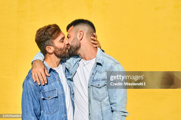 gay couple with arm around kissing each other while standing against yellow wall - bisous photos et images de collection