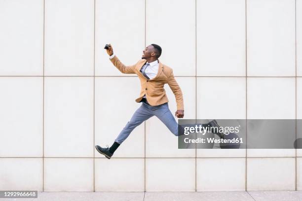 young businessman jumping and taking a selfie in front of a wall - beige jacket stock pictures, royalty-free photos & images