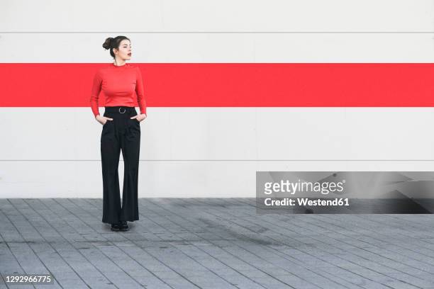 young woman with hands in pockets looking away while standing against wall - coolpad stockfoto's en -beelden